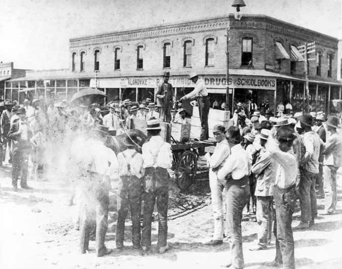 Two men on wagon with display and crowd observing, brick drugstore in background