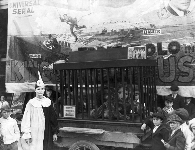 White male clown and costumed lion performer in cage cart with audience boys and circus banner
