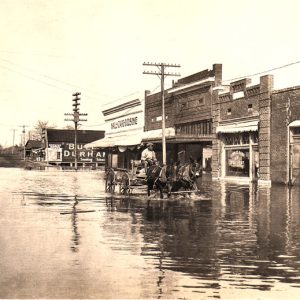 Black man drives two mules and wagon through flooded city street, brick buildings, large sign for "Bull Durham Smoking Tobacco"