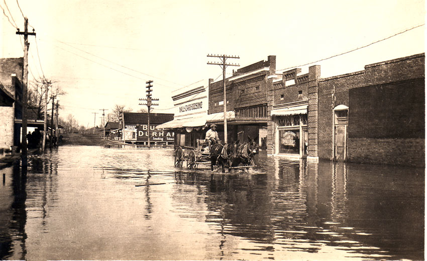 Black man drives two mules and wagon through flooded city street, brick buildings, large sign for "Bull Durham Smoking Tobacco"