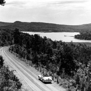 Convertible heads toward mountain lake on tree-lined gravel road, dock in distance