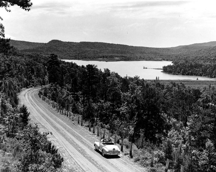 Convertible heads toward mountain lake on tree-lined gravel road, dock in distance