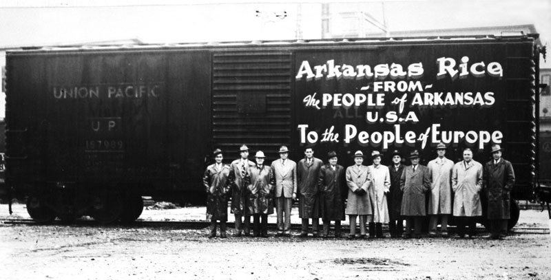 Group photo white men in suits, hats, stand by railcar marked "Union Pacific" with painted sign "Arkansas Rice from the people of Arkansas U.S.A. to the people of Europe."