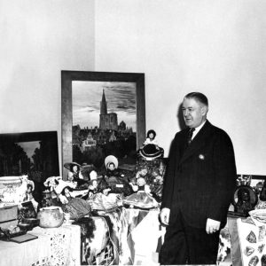 White man in suit, tie, standing among tables with various items, paintings, dolls, tableware