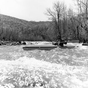White man in kayak holding paddle across chest with river rapids, mountain ridge