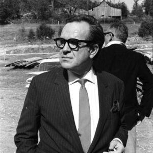 White man in suit and glasses holding a cigarette in front of another white man, in rural setting with barn.