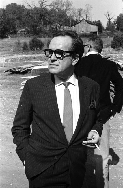 White man in suit and glasses holding a cigarette in front of another white man, in rural setting with barn.
