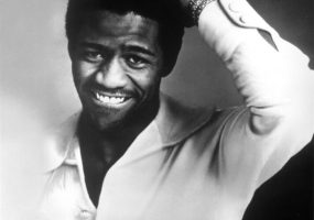 African-American man in button-up shirt smiling with his left hand on his head