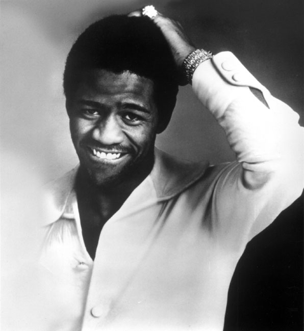 African-American man in button-up shirt smiling with his left hand on his head