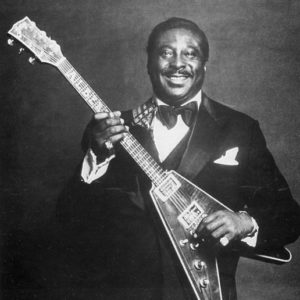 Portrait black man smiling in tuxedo posing with wood veneer triangular body electric guitar