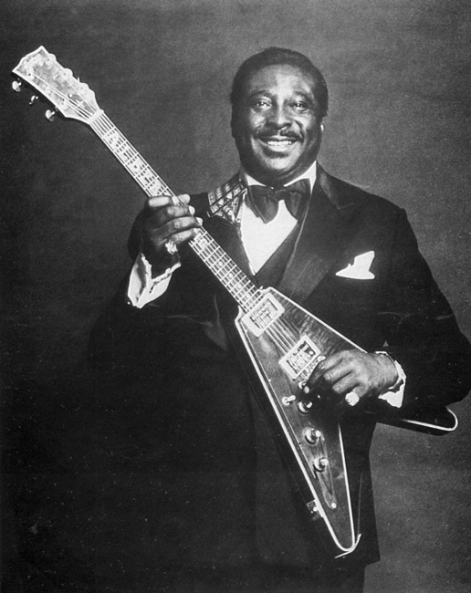 Portrait black man smiling in tuxedo posing with wood veneer triangular body electric guitar