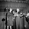 Old white man playing a fiddle on stage with old white woman and white guitarists