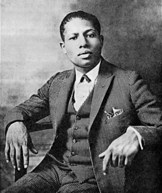 Portrait photo of a seated young black man with a serious expression wearing a three-piece pinstriped suit bow tie and pocket square