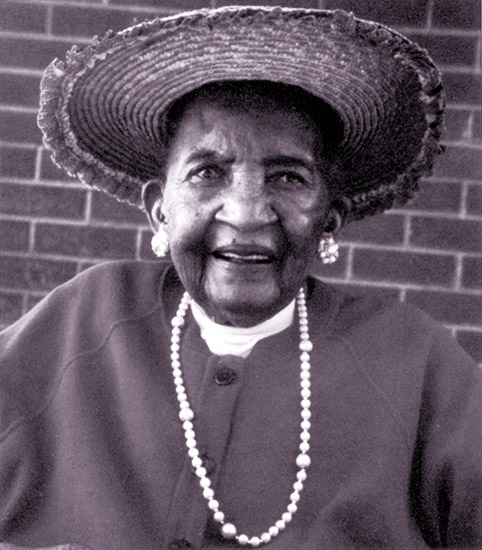 Old African-American woman smiling in hat and necklace with earrings sitting with brick wall behind her