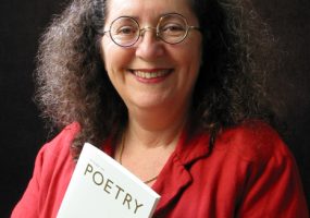 White woman smiling round glasses curly hair red jacket necklace holding booklet titled "Poetry"
