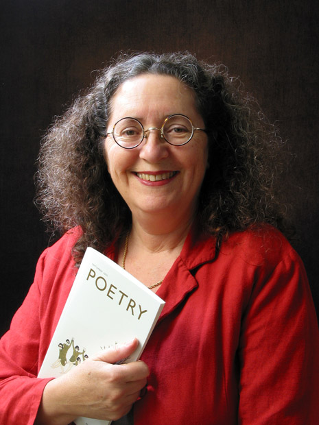White woman smiling round glasses curly hair red jacket necklace holding booklet titled "Poetry"