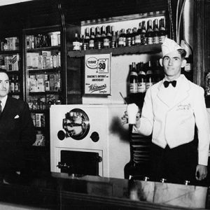 White man in suit and white man in uniform with ice cream in his hand standing behind counter in store