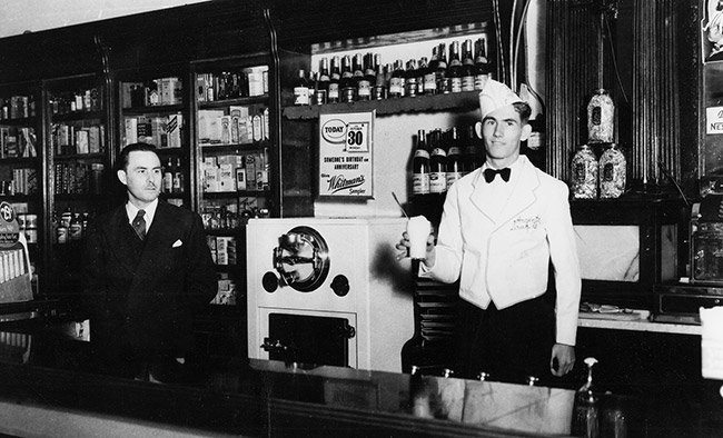 White man in suit and white man in uniform with ice cream in his hand standing behind counter in store
