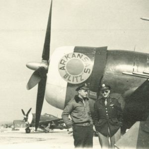 Two men in air force uniforms on an airfield, standing by the nose of an airplane on which is painted "Arkansas 'Blitz'"