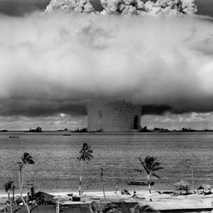 Massive explosion and mushroom cloud over water as seen from an atoll, with various ships dimly visible near the base of the mushroom cloud