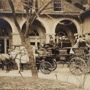 White men in suits one woman pose in horse drawn carriages by arched brick building