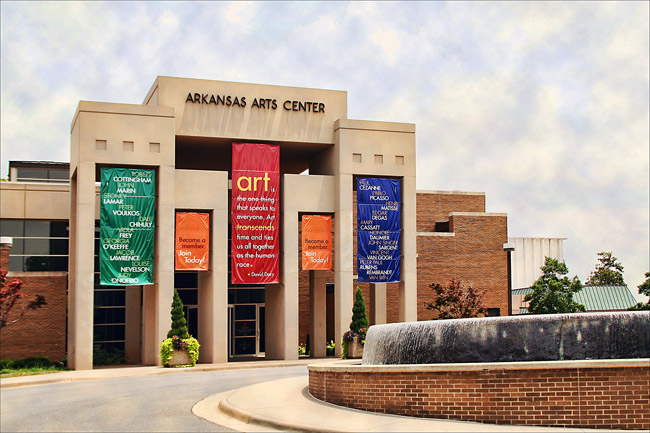 Multistory brick and concrete building with "Arkansas Arts Center" on front with vertical banners circular driveway and large fountain