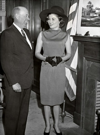 White man and woman stand smiling in formal attire by fireplace and U S flag