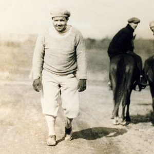 White man in hat, sweater, baseball pants, walking away from two men on horses in background