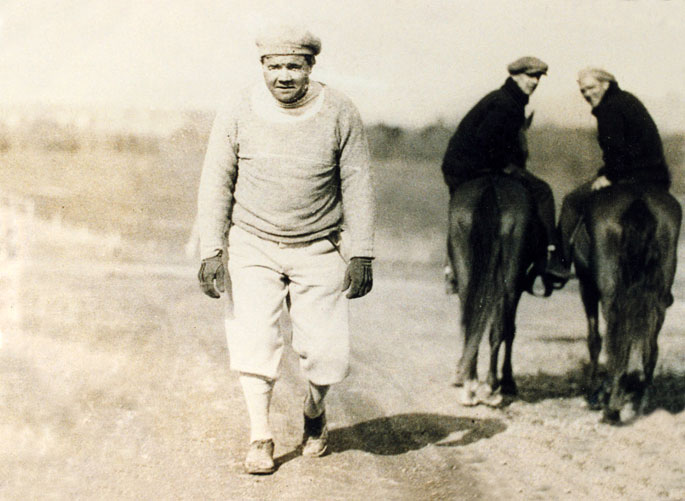 White man in hat, sweater, baseball pants, walking away from two men on horses in background