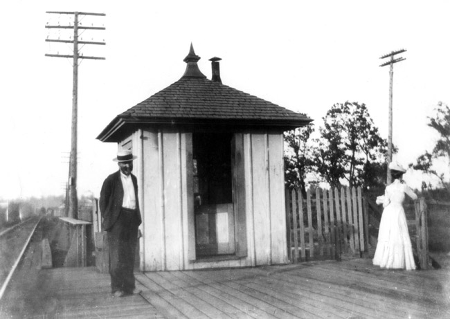 man in straw hat outside small building with pointed roof standing alongside railroad tracks