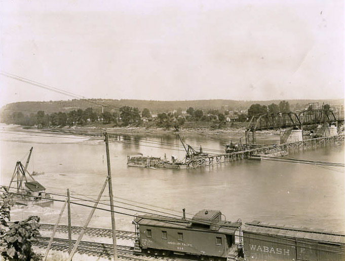 Steel arch bridge being built over a river with train cars on tracks in the foreground