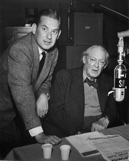 White man in suit talking with older white man sitting with microphone in the foreground