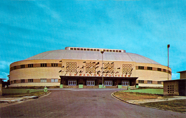 Multistory rounded concrete building with "T H Barton Coliseum" on the front with four entrances and front-mounted flag poles