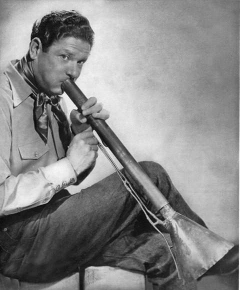 white man dressed as cowboy blowing into long, tubular musical instrument