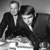 Two white men in suits one blows out "A" shaped cake candles near Alabama elephant figurine
