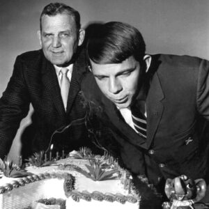 Two white men in suits one blows out "A" shaped cake candles near Alabama elephant figurine