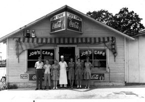 White man in chef whites posing outside cafe with five women and a man under "coca-cola" sign