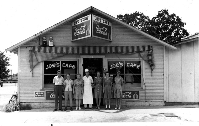 White man in chef whites posing outside cafe with five women and a man under "coca-cola" sign