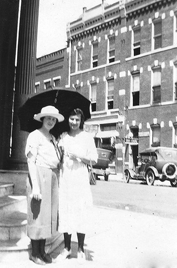 Two white women in dresses on street corner with umbrella and cars and buildings behind them
