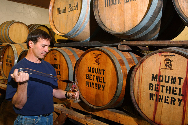 White man sampling wine from aged wooden barrels labeled "Mount Bethel Winery"