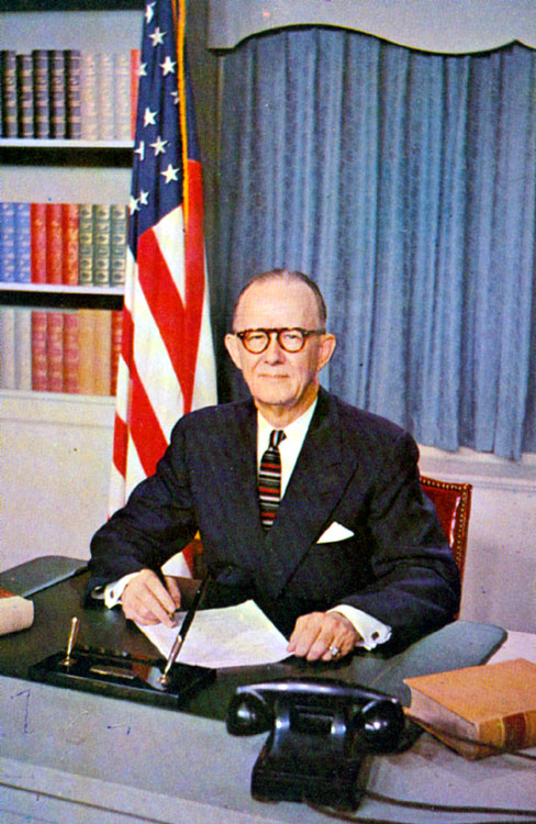 Old white man in suit and tie with at his desk with curtains, flag, and bookshelves behind him