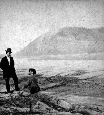 two white men in hats, one standing and another sitting along the edge of a river with large hill in background