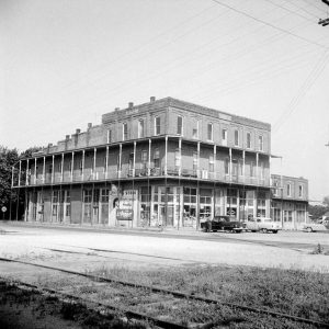 Three story brick building of shops with long balconies with cars parked by it and railroad tracks in foreground
