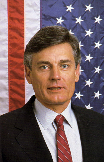 white man in suit and tie with American flag backdrop