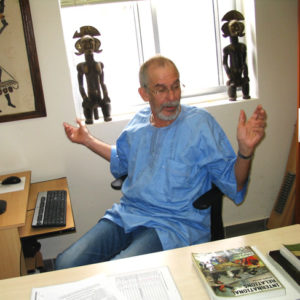 Old white man with glasses and beard in blue shirt and jeans sitting at computer desk in office decorated with African artwork