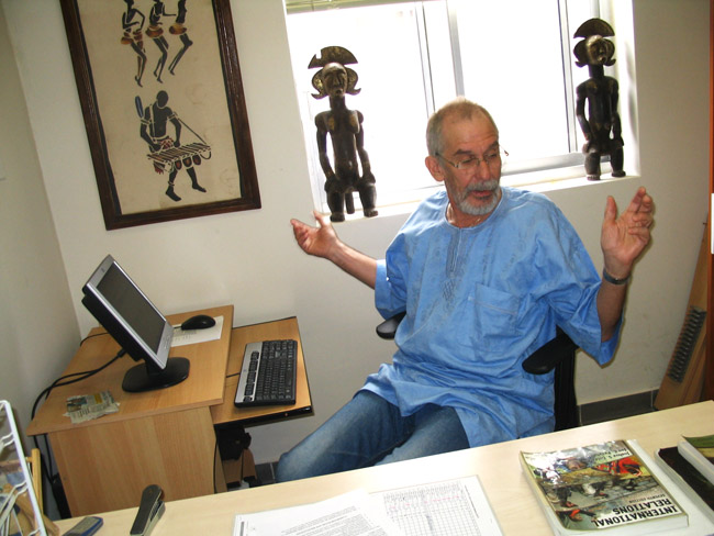 Old white man with glasses and beard in blue shirt and jeans sitting at computer desk in office decorated with African artwork