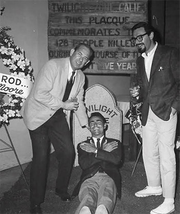 White man in suit bending over to white man laughing in front of fake tombstone next to tall white man in glasses