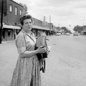 White woman carrying a large bag across the street with storefronts in the background.
