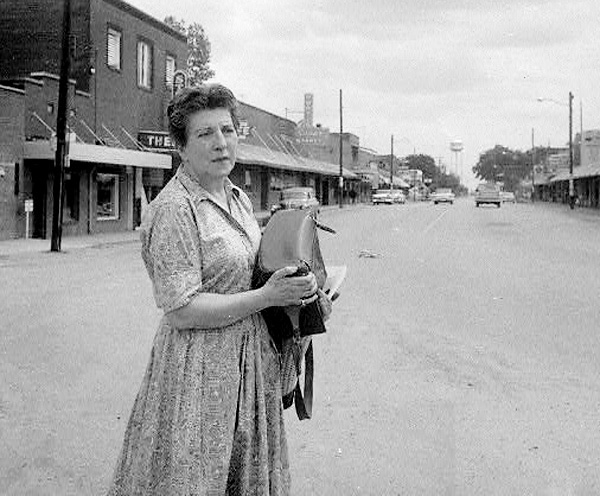 White woman carrying a large bag across the street with storefronts in the background.