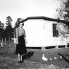 Young white woman in dress and chickens standing outside house with fence and outbuilding